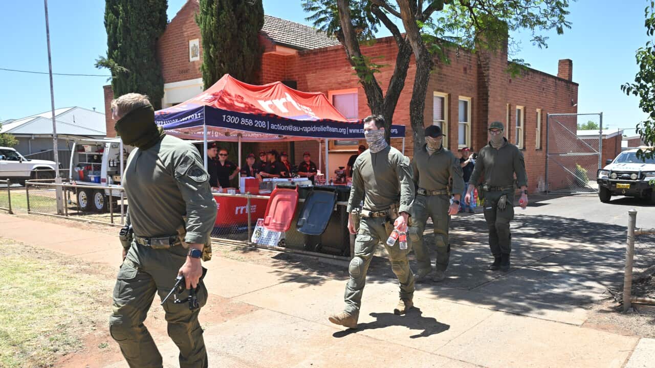 Four men in olive-green tactical gear and face gaiters walk along a paved path in front of a red brick building and a Rapid Relief Team tent.