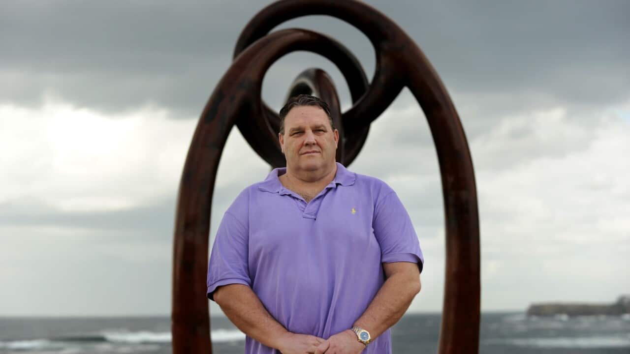 A man in a purple polo stands in front of a metal sculpture by the ocean.