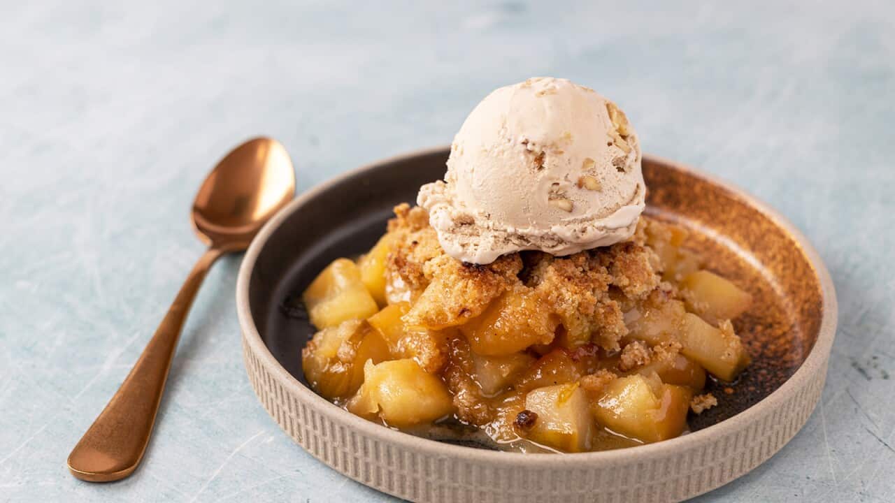 A serve of juicy apple crumble sits in a low round serving dish, with a scoop of ice-cream on the top. A copper-coloured spoon sits alongside the dish.