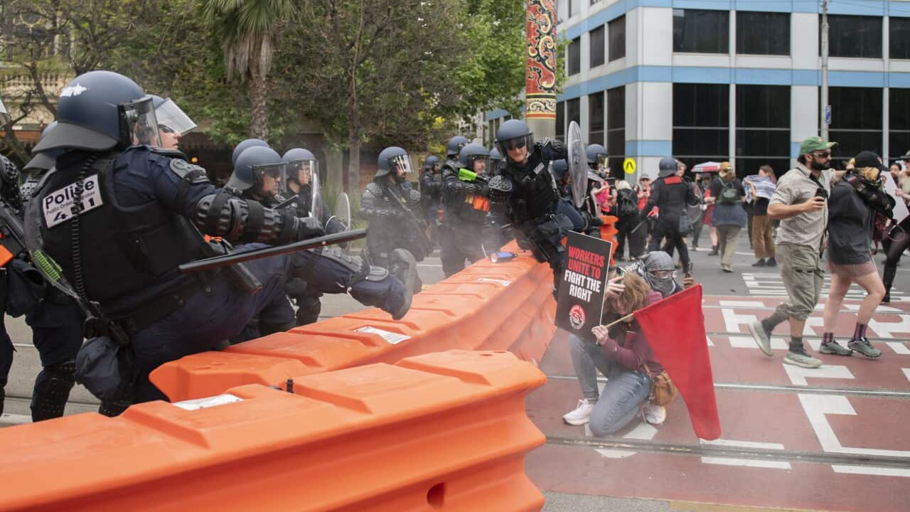Police wearing riot gear standing behind an orange road barrier while protesters crouch on the other side