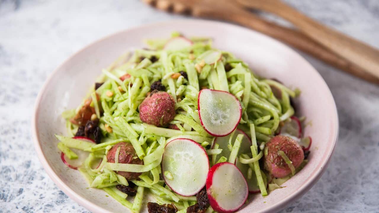 A green-hued salad sits in a round pale pink bowl. Wooden salad servers sit behind the bowl, to one side.