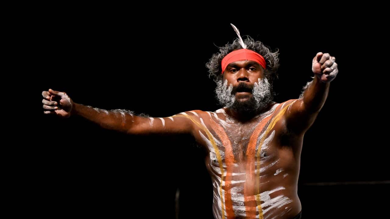 An Indigenous man wearing a red head band and with paint on his body dances with his arms extended in front of him