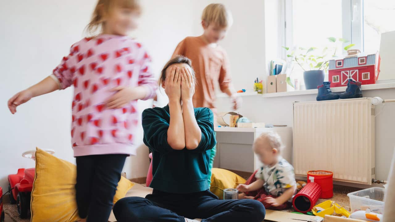 A mother sits on the floor in the middle of a room, her face in her hands, three children running around her.