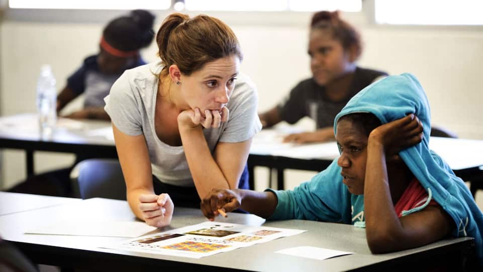 A teacher leans down on the desk to listen to a student next to them.