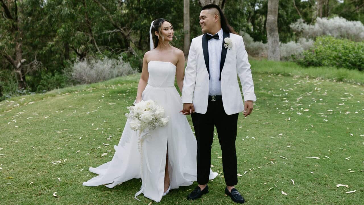 A woman in a white wedding dress, holding hands with a man in a white suit, standing on grass in front of a large tree.