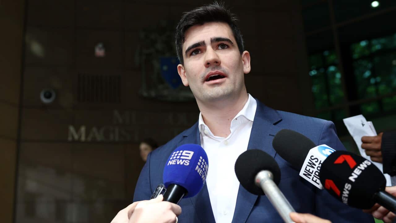 A man in a blue suit and dark hair stands in front of microphones