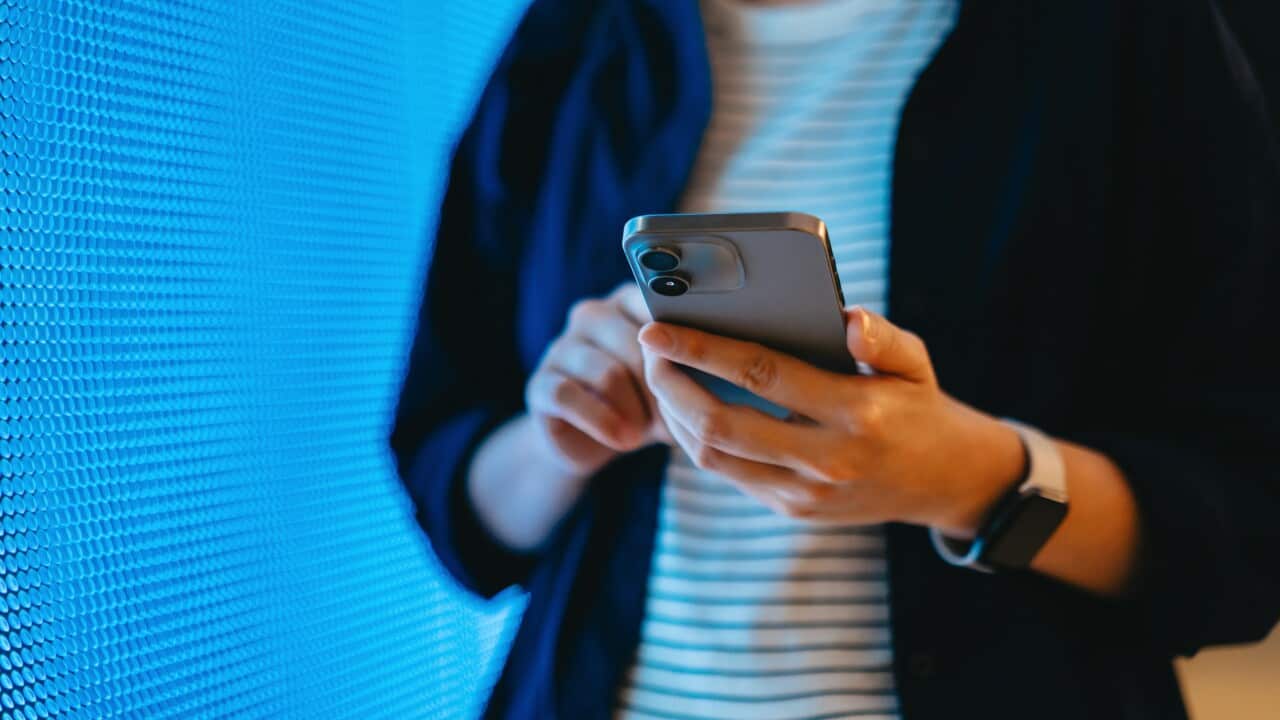 Mid-section of a person using smartphone against blue coloured illuminated LED digital display screen in the dark.