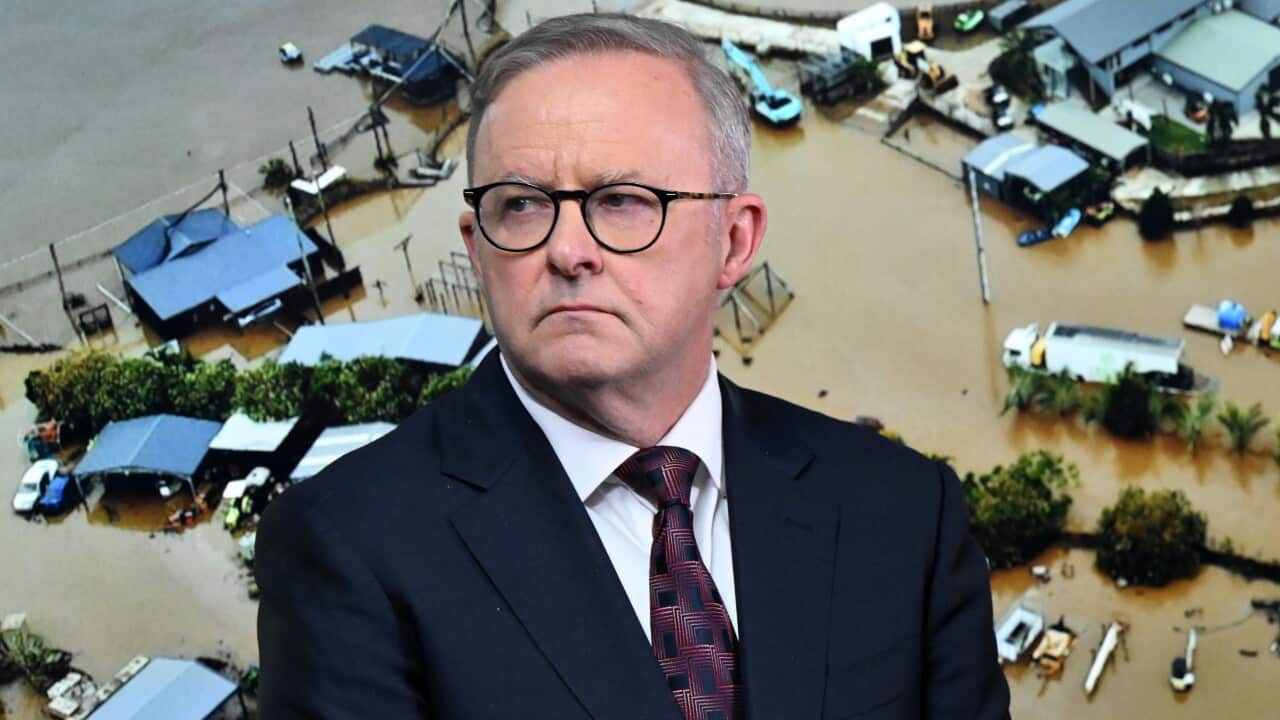 A man wearing a suit, tie, and glasses standing in front of a screen that shows a flooded scene.