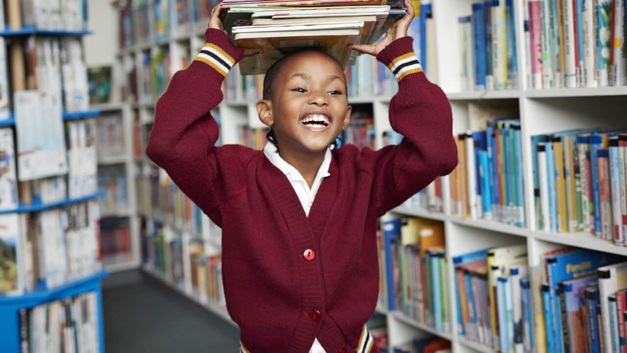 Cute schoolgirl smiling & balancing stack of books on the head at library