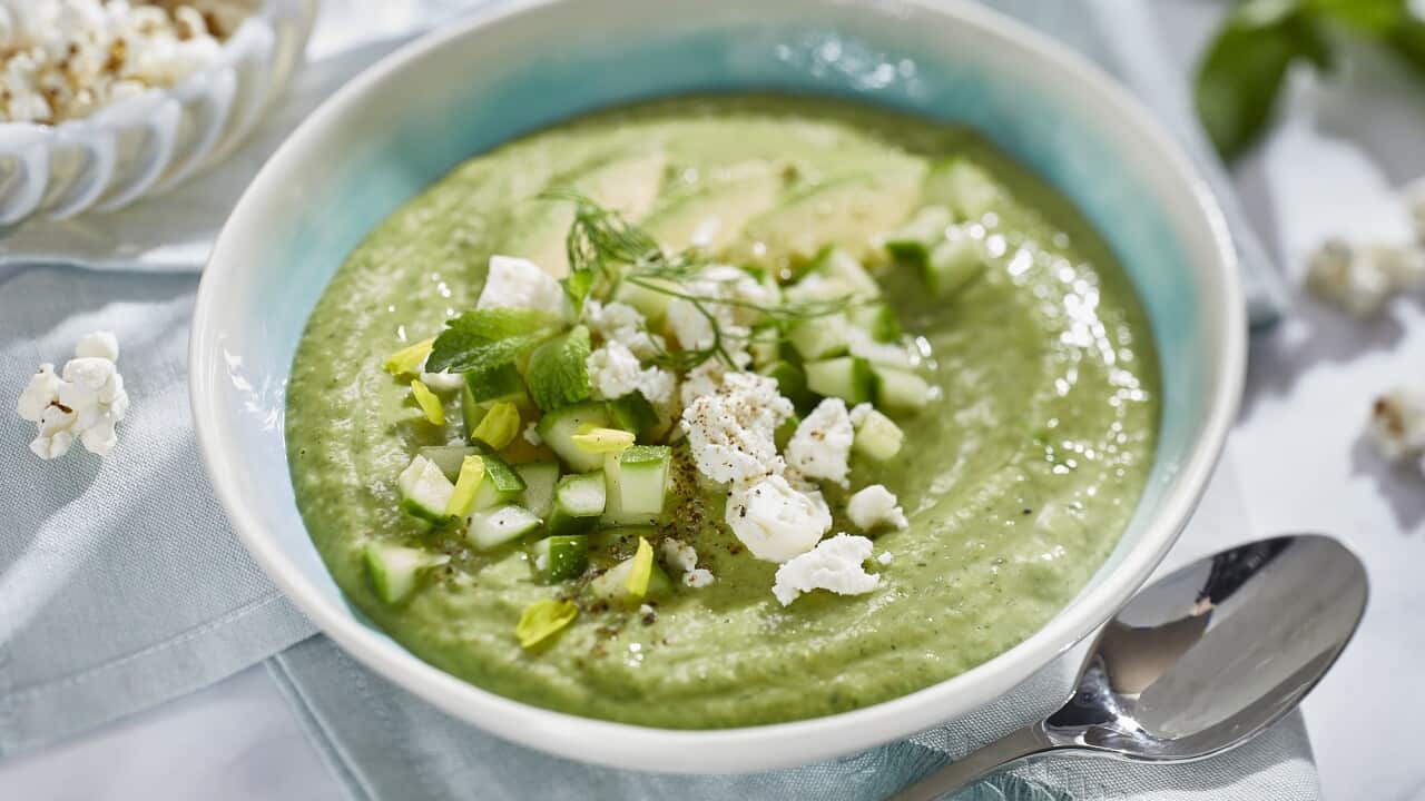 A white bowl with a pale-blue glazing showing on the inside holds a thick green soup, topped with a tumble of feta. avocado and cucumber. The bowl sits on a pale blue napkin, and a silver spoon sits alongside the bowl.