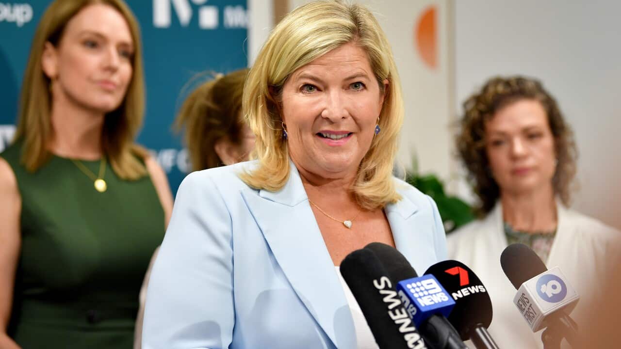 A woman wearing a blue blazer and white top speaking while standing in front of microphones.