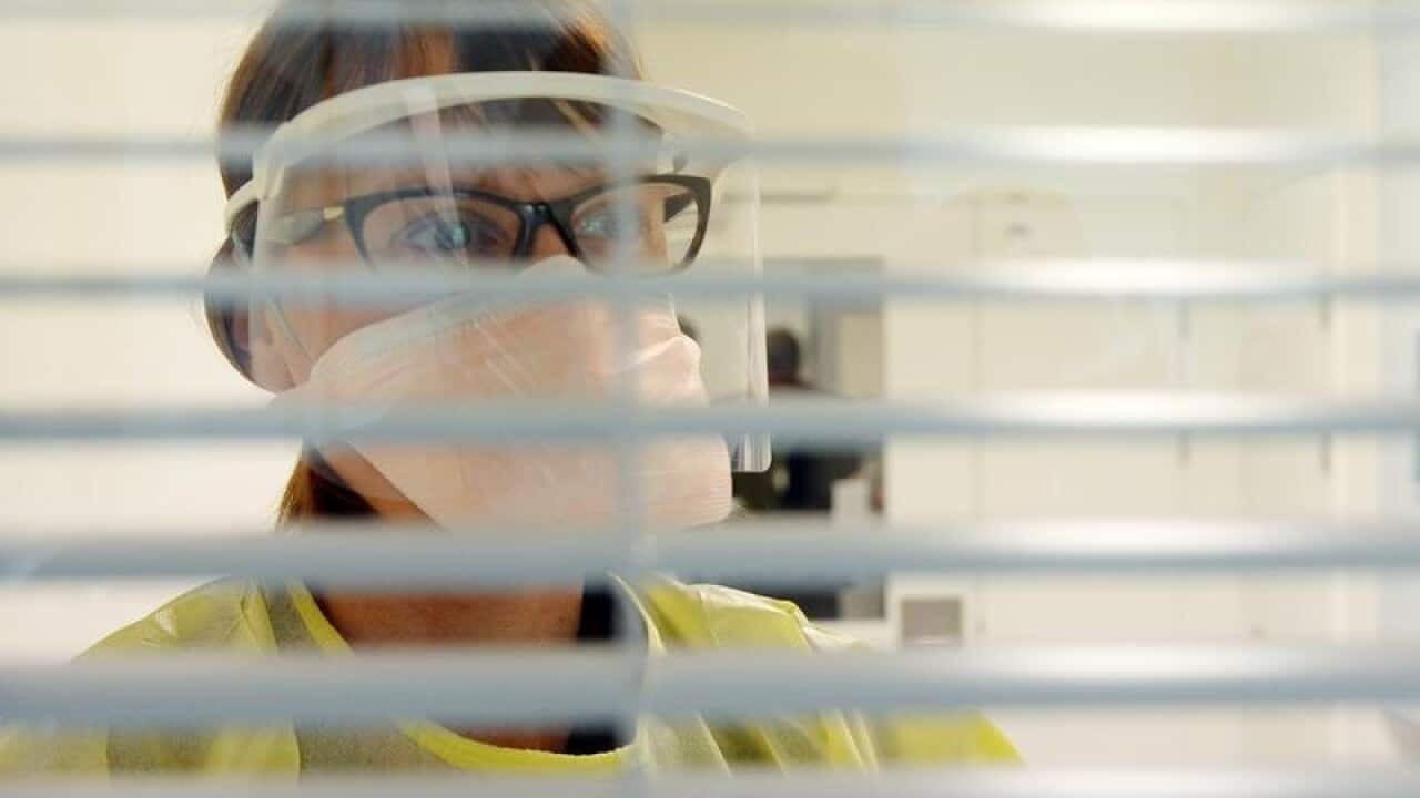 A nurse in protective gear looks through into an isolation room