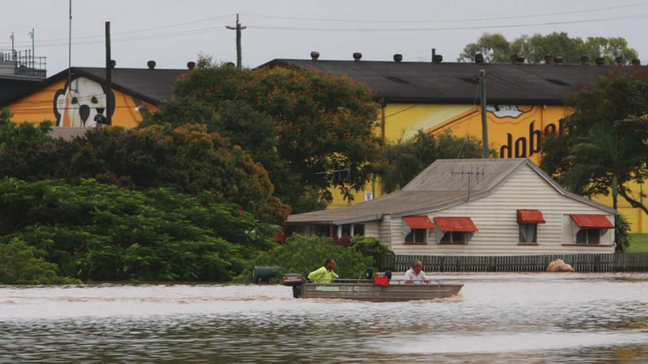 A small tin motorboat crosses a flooded street in the city of Bundaberg, Queensland. Two men sit inside the boat, one in high vis yellow, the other in a white shirt. Behind them is a white house with a tin roof and red eaves, the water half way up its outer fence. Behind the house, the famous Bundaberg Rum factory.