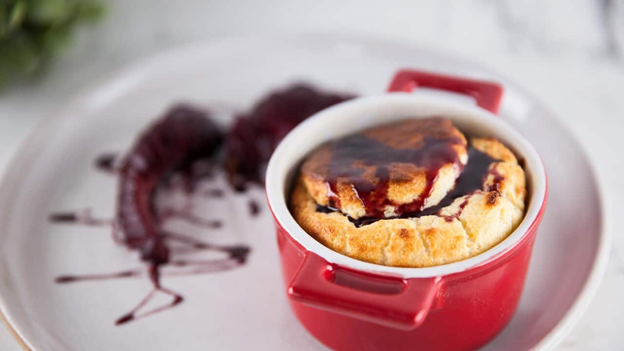 A small red ramekin, holding a souffle, sits on a white plate. Two dark pear pieces sit on the plate beside the ramekin, in a drizzle of dark sauce.