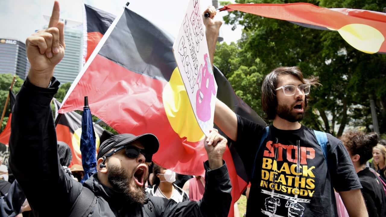 Protesters hold Australian Aboriginal flags and signs, including one reading "Stop Black Deaths in Custody," while shouting during a demonstration.