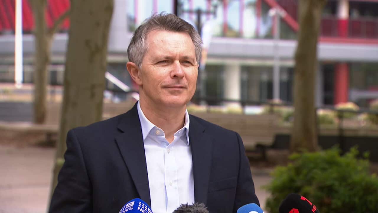 A man in a black suit with a light blue shirt, with trees in the background and microphones in front of him.