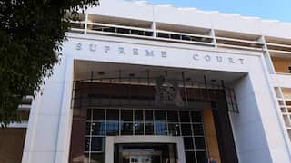 A low-angle exterior shot of a modern, white building with the words "SUPREME COURT" in silver lettering above the entrance, with a tree branch partially obscuring the left side.