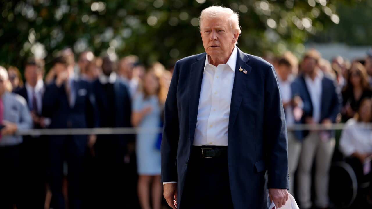 A close-up photograph features Donald Trump walking outdoors, dressed in a navy suit jacket, dark trousers, and an unbuttoned white shirt, holding what appears to be a white hat in his right hand.