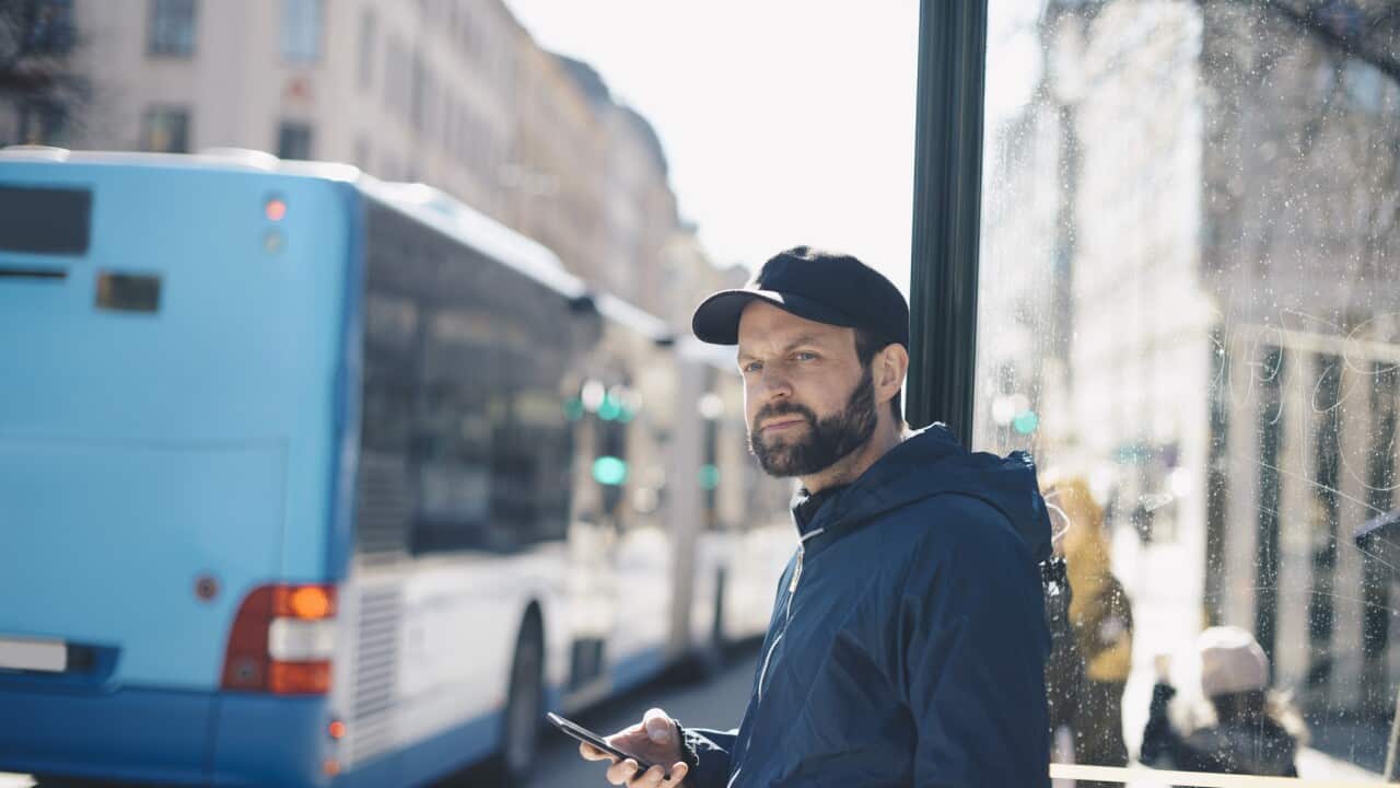 Thoughtful man holding mobile phone while leaning on pole against bus in city
