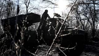 A soldier stands atop a truck-mounted multiple rocket launcher system positioned in a wooded area, silhouetted against a bright, low-hanging sun.