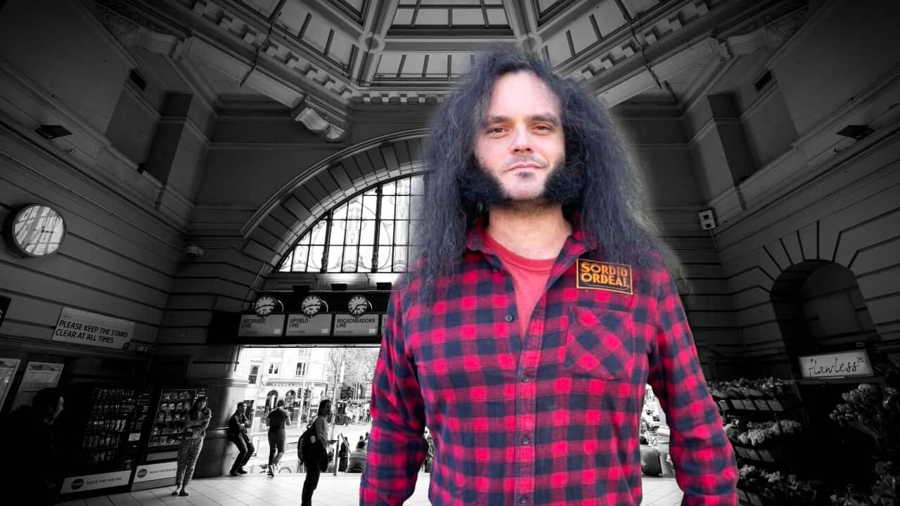 A man with long, curly hair in a red checked shirt with a Sordid Ordeal patch. Behind him is the entrance of a grand old train station