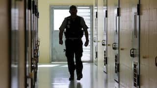 A male prison guard in uniform walks down a corridor which is lined with doors to prison cells.