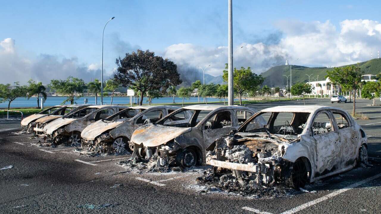 A row of burned-out cars in a parking lot, with a view of the sea and green hill behind them.