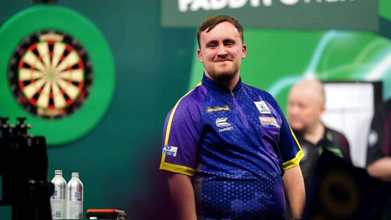 A young man wearing a shiny polo shirt stands smiling in front of a dart board.