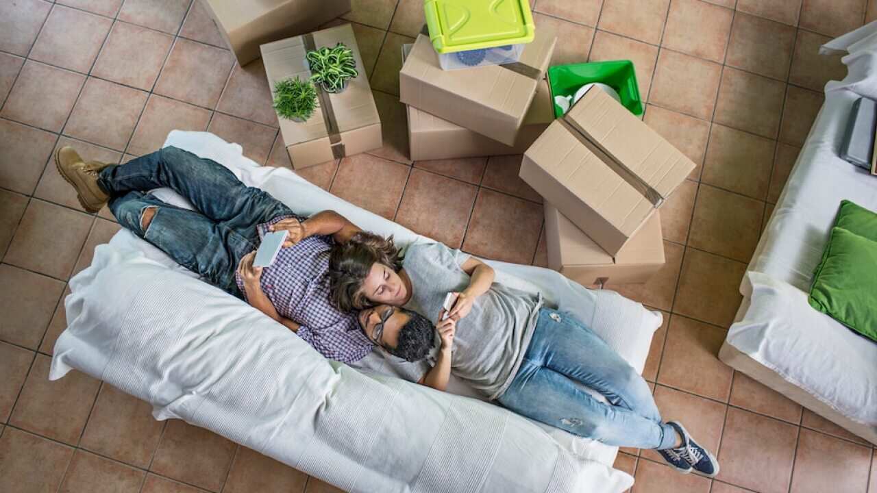 Couple checking their own digital devices on the social networks on the sofa with all the boxes to unpack at the new home.