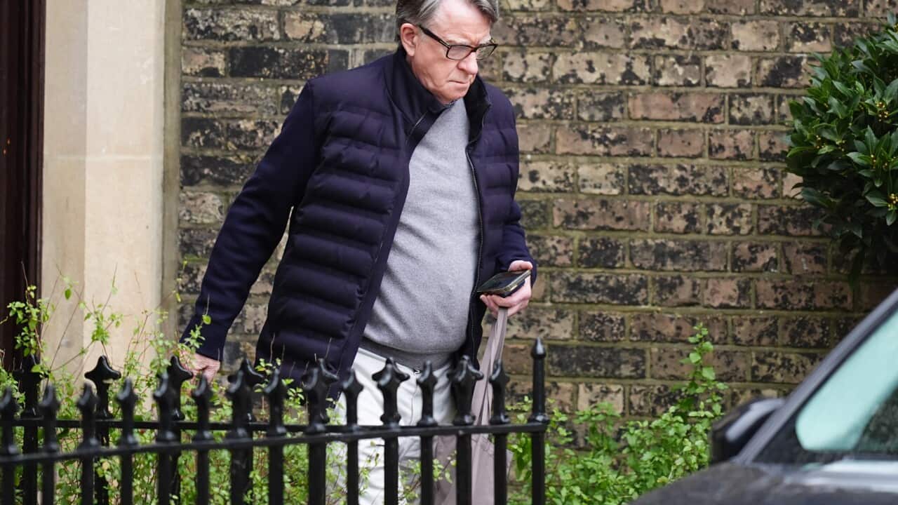 An older man wearing glasses and a dark jacket walks past a brick wall while carrying a folded umbrella and phone.