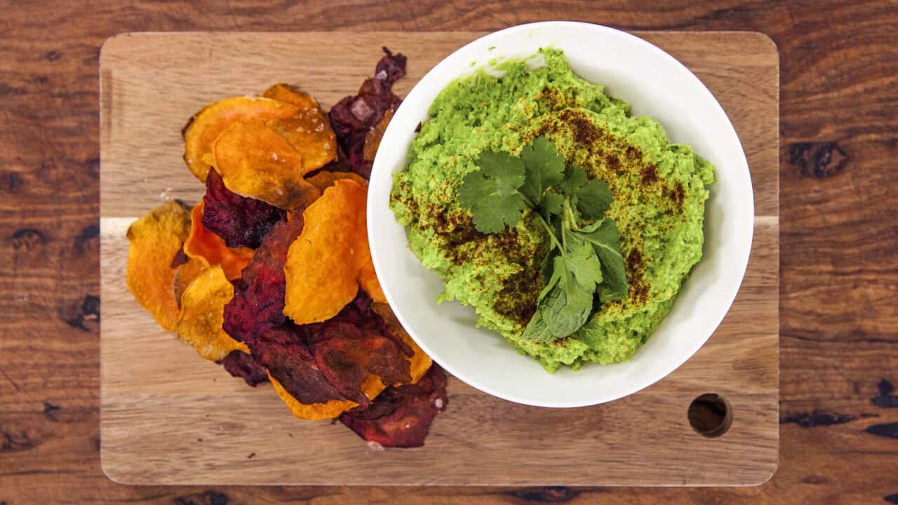 Seen from overhead, a white bowl filled with a green dip sits on a wooden board, alongside a pile of vegetable crisps.