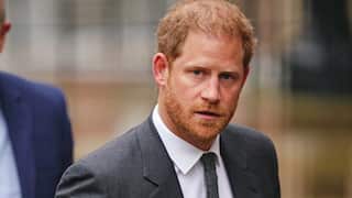 Man with red hair and beard wearing a grey suit is framed from the shoulders up, looks straight to camera as he walks outside.