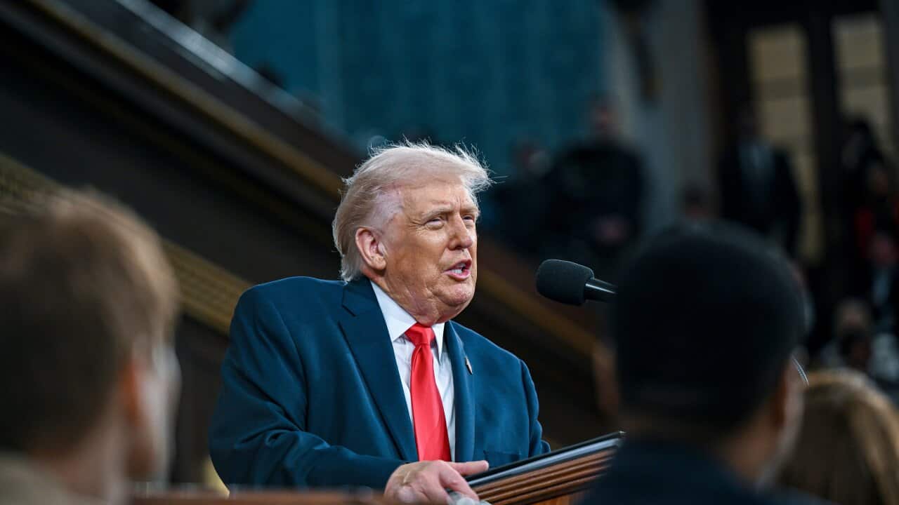 Donald Trump, standing at a podium and speaking into a microphone. The backs of other people's heads are visible in front of him.