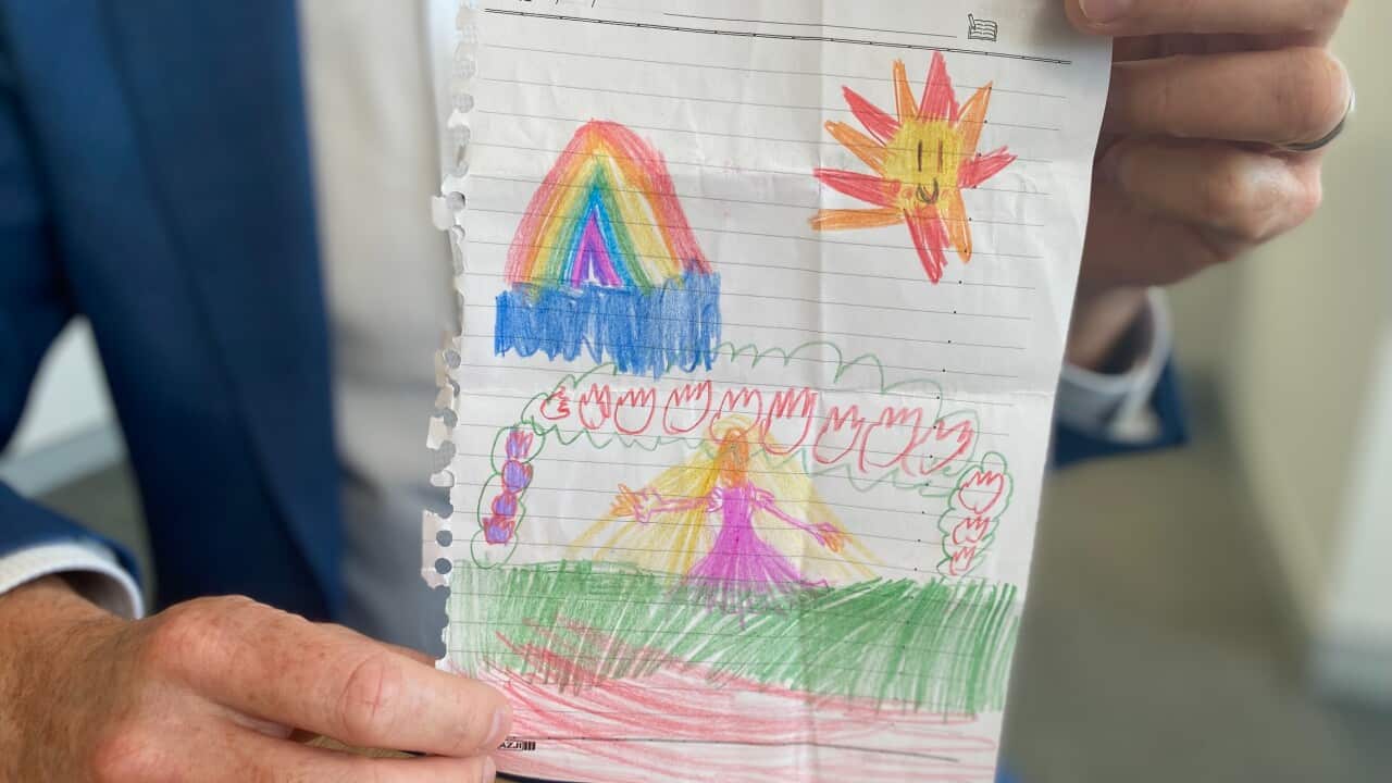 A pair of hands, belonging to a person in a blue blazer, holds up a child's drawing on a piece of torn, lined paper. The artwork features a brightly coloured rainbow, a smiling sun, and a figure in a pink dress surrounded by a fence of colourful, flame-like shapes, all drawn with crayons.