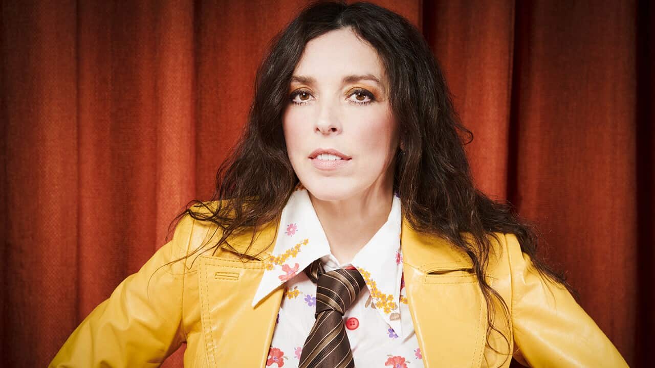 A womain in white and floral shirt, brown striped tie and jellow jacket, with long brown hair, looks at the camera.
