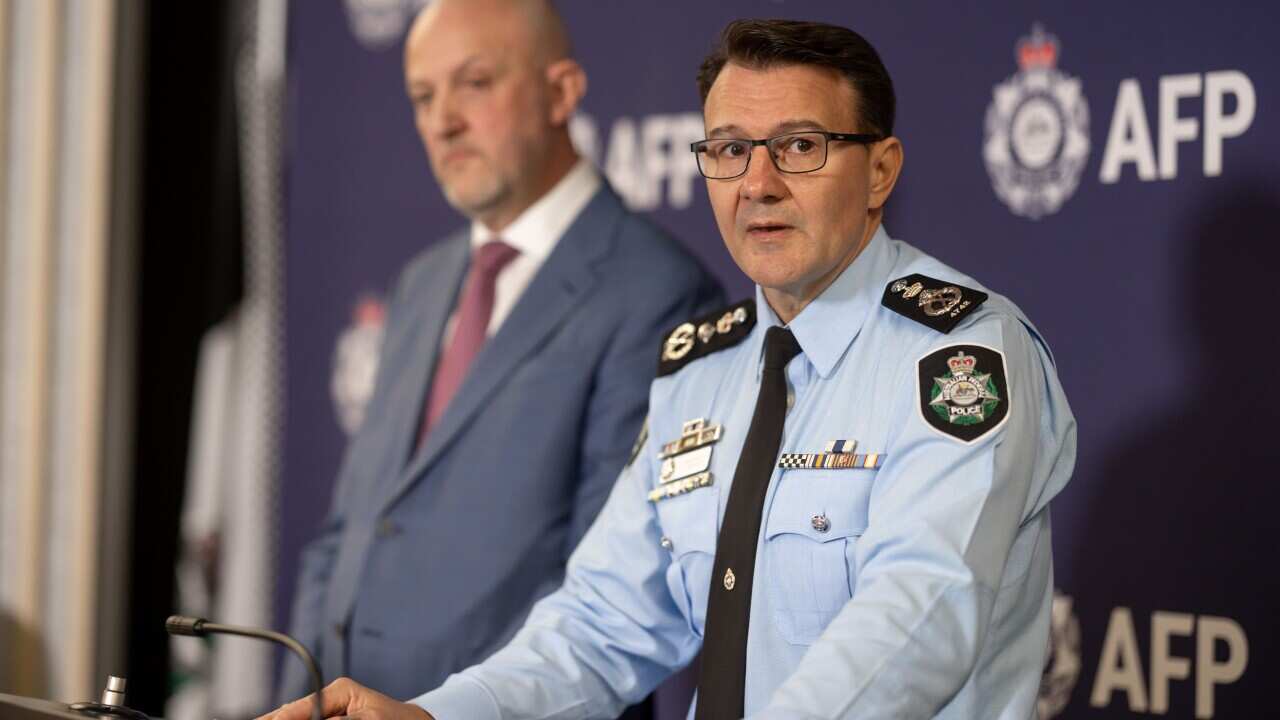 Australian Federal Police Commissioner Reece Kershaw speaking while standing at a lectern. Standing next to him at ASIO Director-General Mike Burgess wearing a suit and tie.