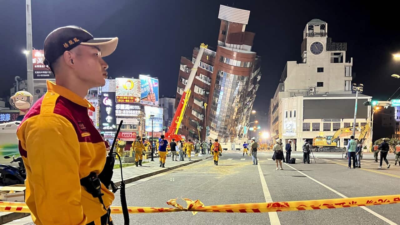 A rescue worker in a bright yellow uniform stands near the cordoned-off site of a leaning building.