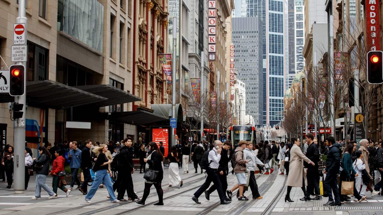 A crowd of people walk across a busy city street with a tram visible in the background