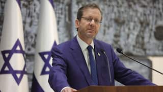 A middle-aged white man wearing a navy suit and glasses before a lectern. Israeli flags hang in the background.