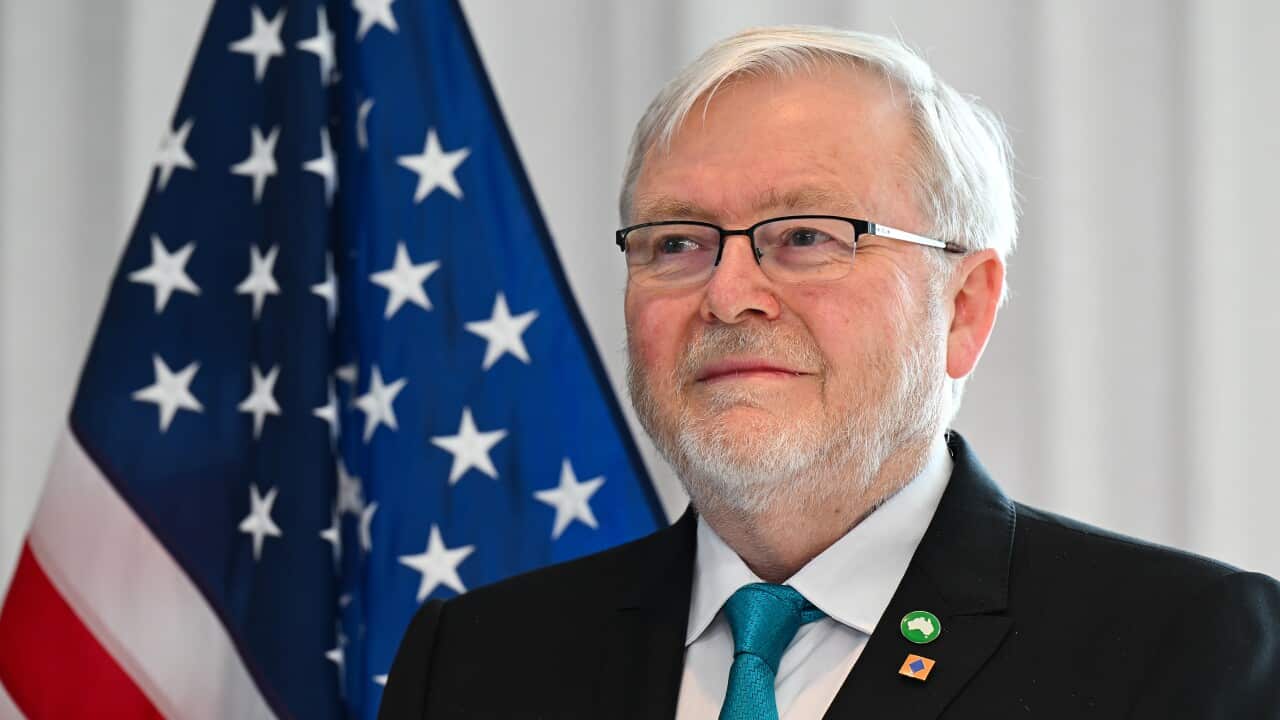 A man with a white beard and hair, wearing a suit and a blue tie, is smiling as he stands next to the Australian flag.