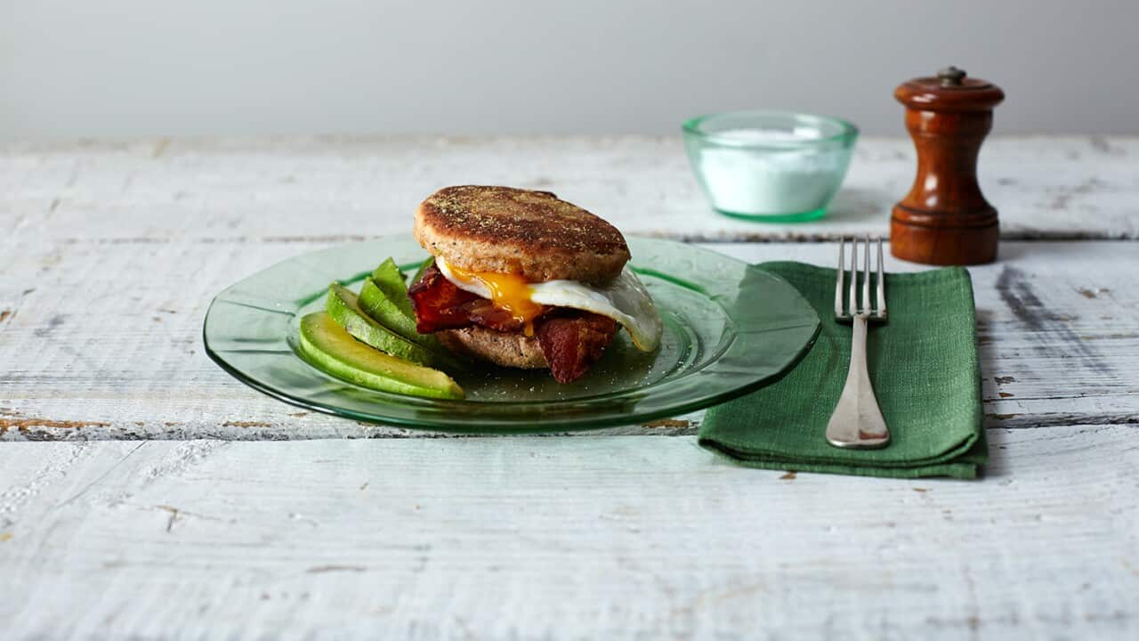 A green glass plate sits on a white-washed wooden surface. On the plate there is a split and filled English muffin, with slices of avocado sitting beside it. Beside the plate, a silver fork sits on a green napkin.