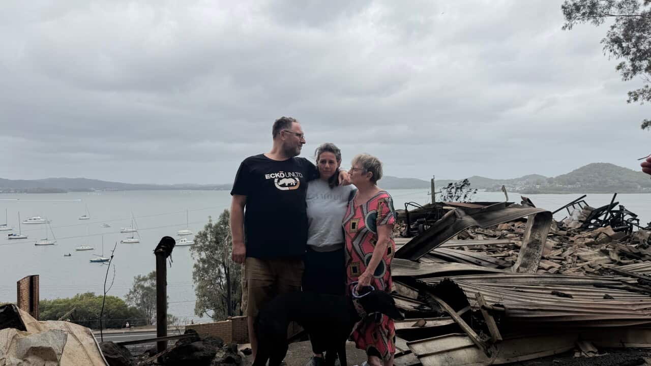 Three people stand solemnly next to a burnt-down home.