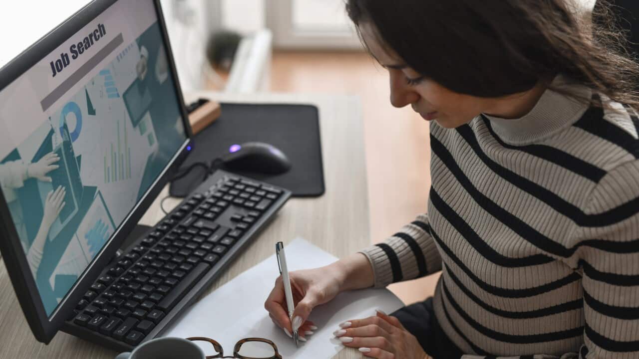 A woman sitting at a desk, writing on a piece of paper. A job search website is open on the computer screen in front of her