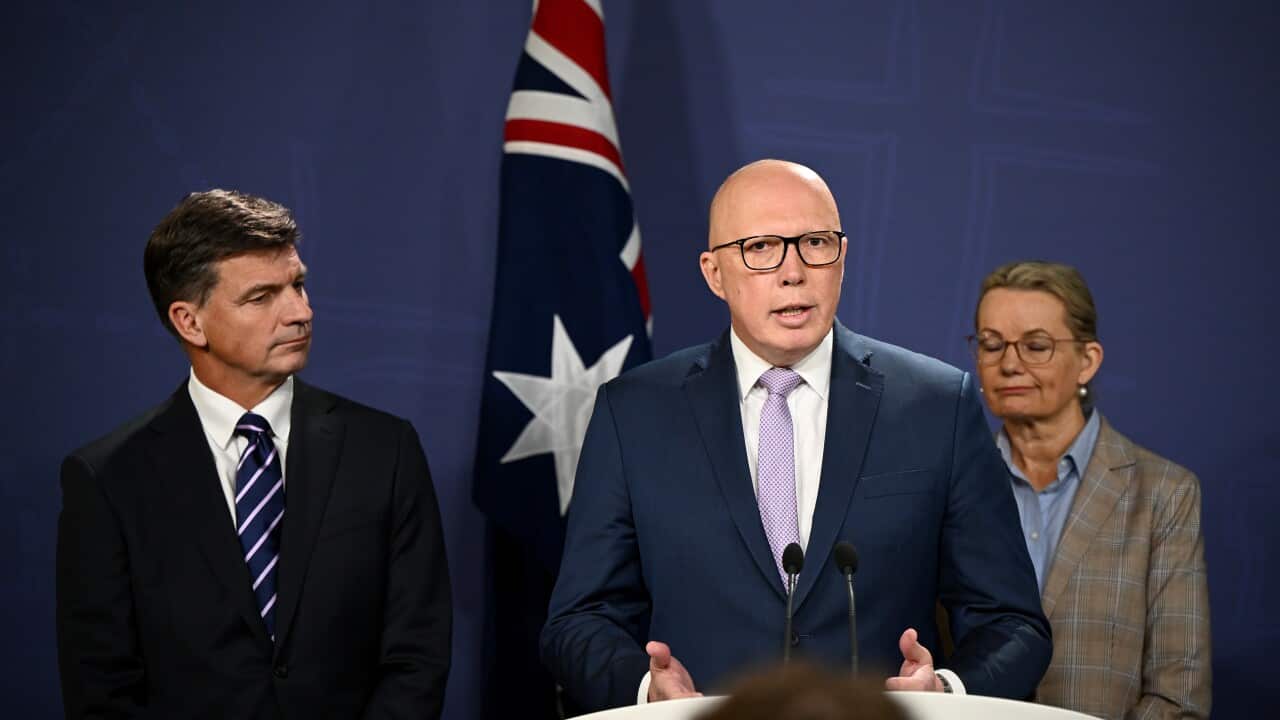 Three people standing, one is behind a podium and speaking into a microphone. An Australian flag is behind them.