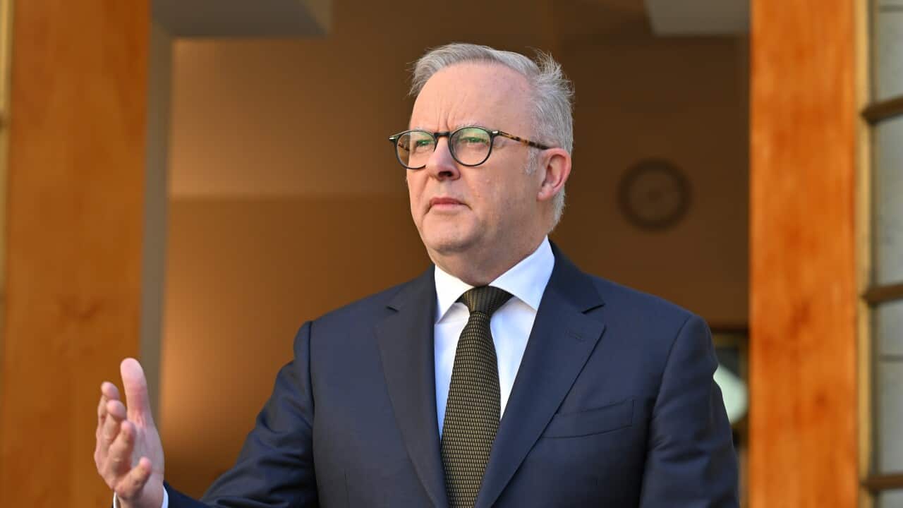 A middle aged white man wearing a dark suit and glasses. He has his right hand in front of him.