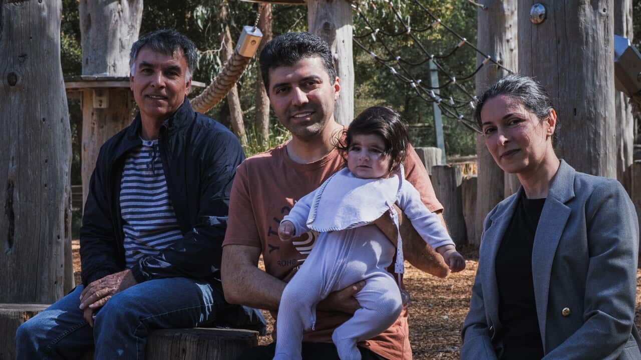 A woman and two men sit on a wooden structure at a playground. A man in the middle is holding a baby.