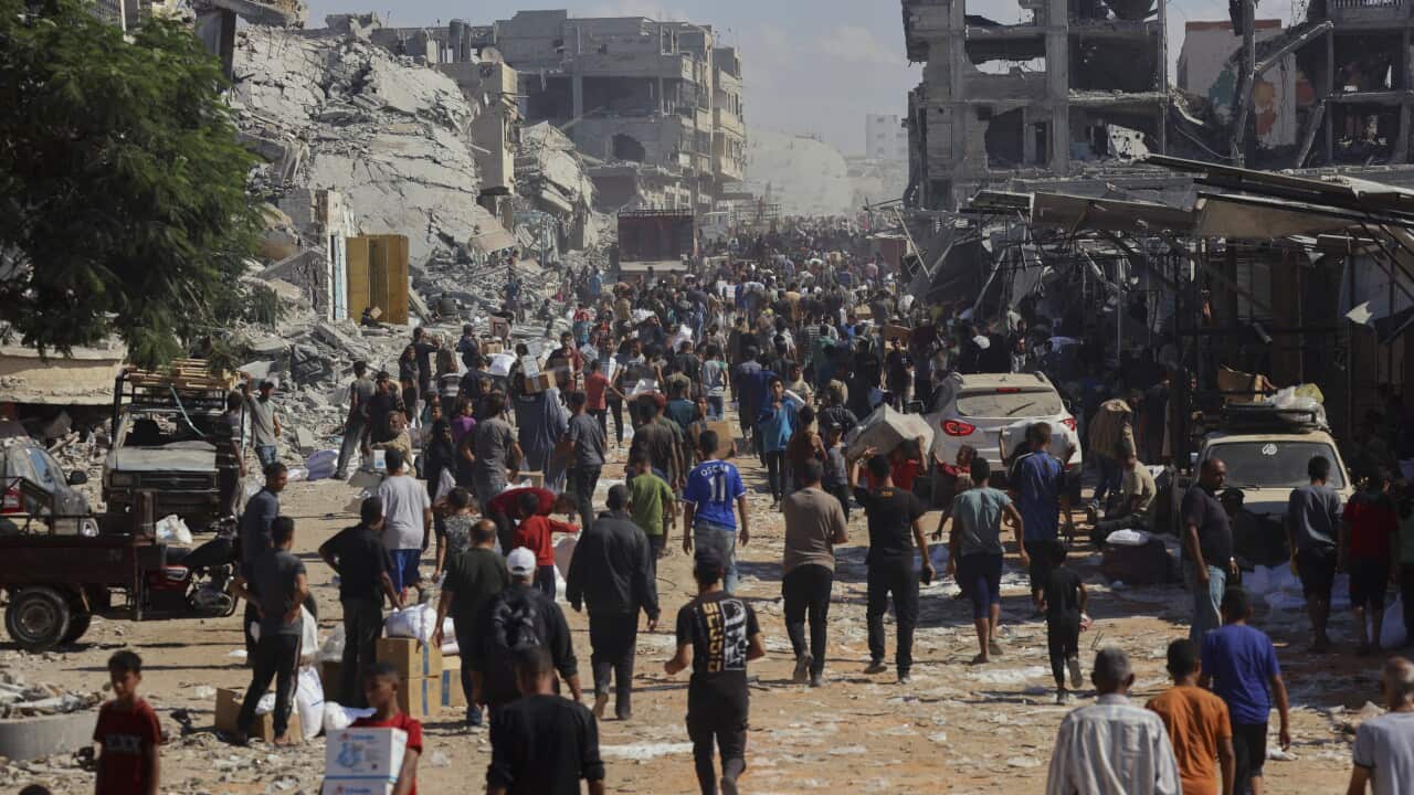 A large group of people walk along a road with heavily damaged buildings on either side.