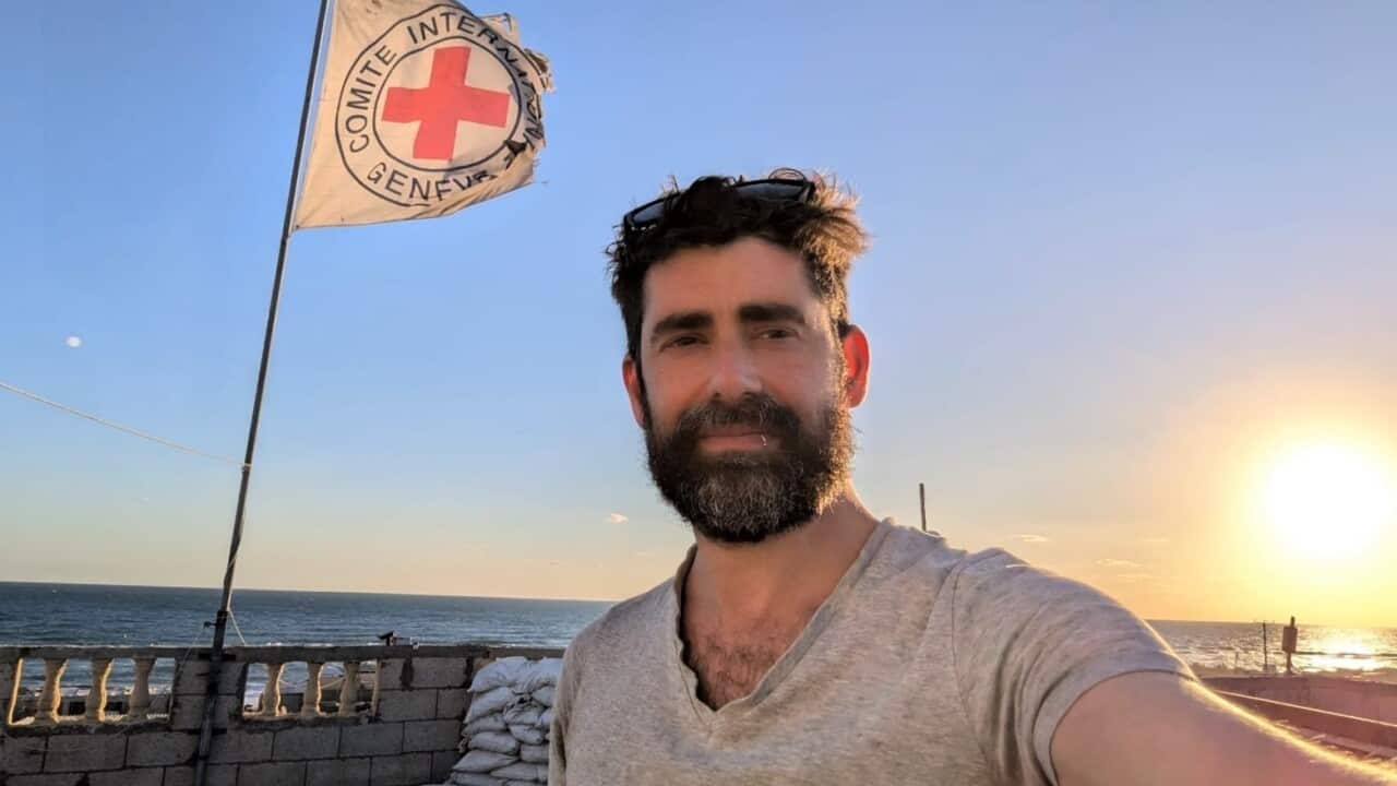 A bearded man in a grey t-shirt stands in front of a Red Cross flag as the sun sets on the horizon.
