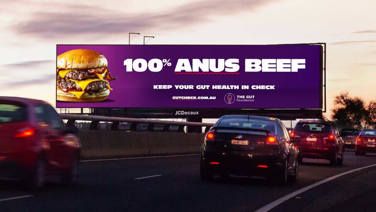 A billboard on a highway that has a hamburger and reads “100 Per Cent Anus Beef“. Cars are driving on the highway in front of it.