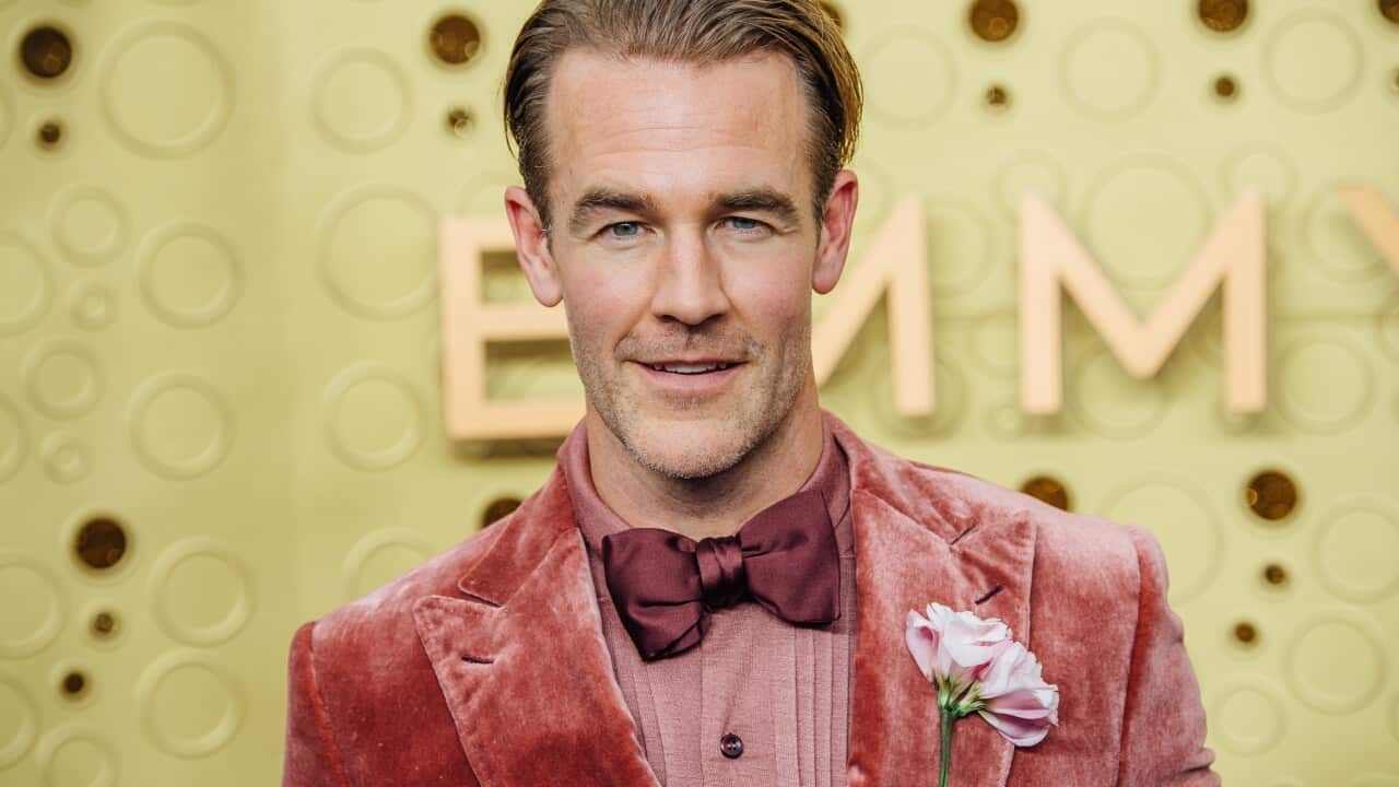 A man wearing a red velvet suit with a pink flower attached to it, in front of a yellow wall.